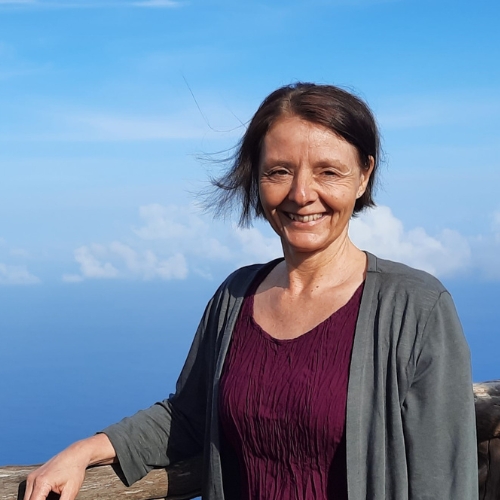Michaela Mischok steht lächelnd an einer Holzbrüstung auf einem Berg mit Blick aufs Meer und die Sonne – Symbol für Ruhe und Gelassenheit